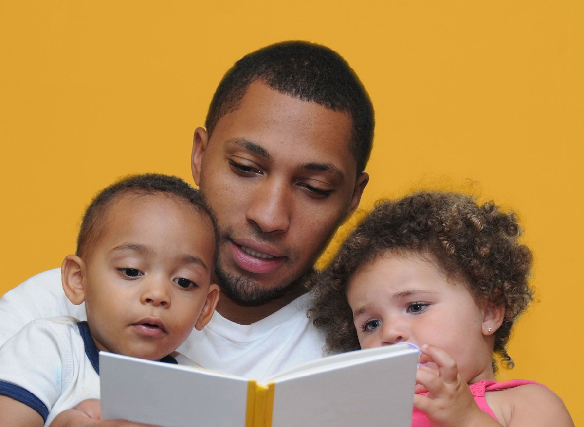 Dad holds and reads to fraternal twins in his lap. Read-aloud with music workshop.