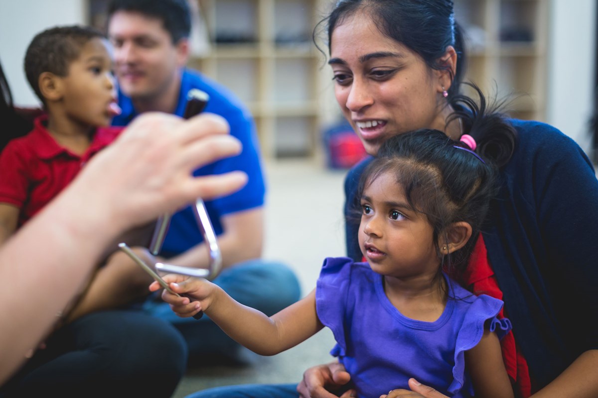 Mother holds 3-year-old daughter in lap while she explores the triangle with her Kindermusik teacher.